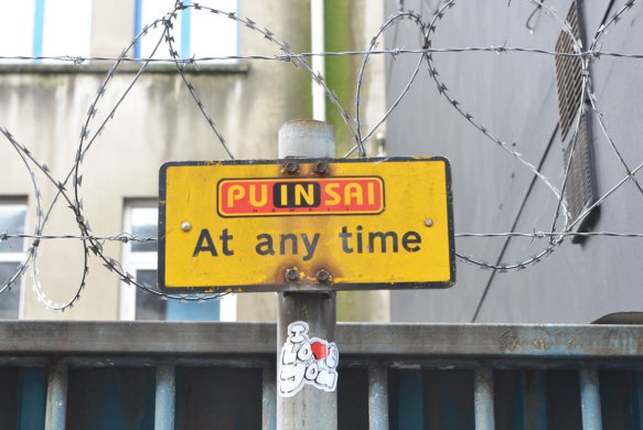 altered sign, now says puinsai Hawaii anytime. yellow sign on a post with barbed sire on either side. Also An 'I love you" sticker on the pole beneath the sign. 