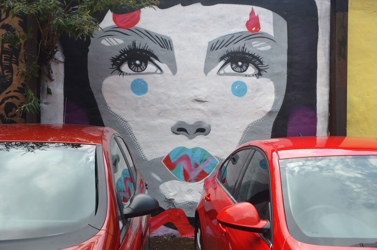 two red cars are parked against a wall on which a mural of a young woman's face is painted. white skin, black hair, grey eyes, 