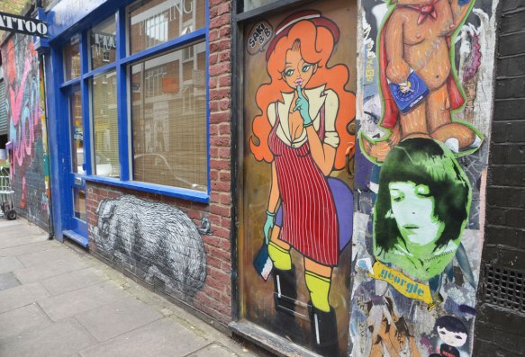 pasteups on a wall, a tall life sized girl by Saki & B, with long orange hair, yellow tights, black boots and a red dress, a woman's portrait in greenish tones by georgie artist, a phoebe, and a black and white pig, 