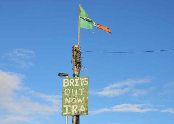 a frayed Irish flag flies on top of a post.  A sign that says Brits out now IRA is also on the post 
