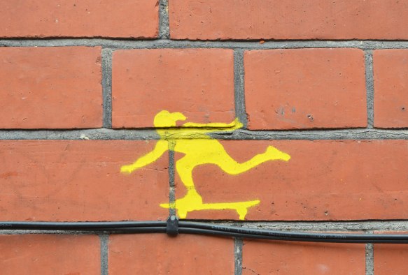 little yellow stencil of a person skateboarding, on a brick wall 