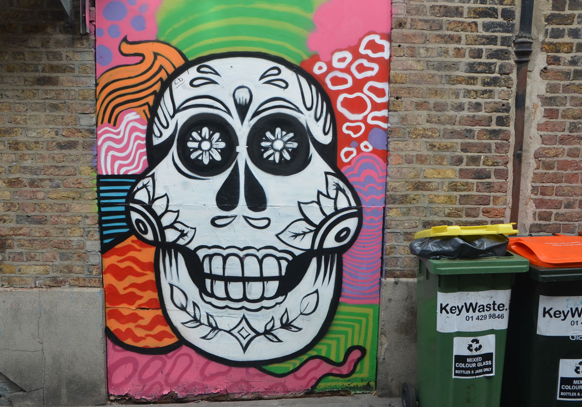a metal street box is covered with a colourful painting of a shite skull decorated like Mexican day of the dead on a many patterened background.