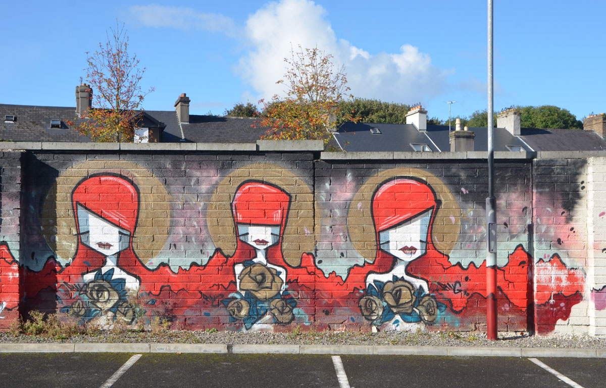 mural on a brick wall in a car park, with houses behind, three stylized women with bright red hair, flowing to their shoulders, all holding bouquets of flowers 