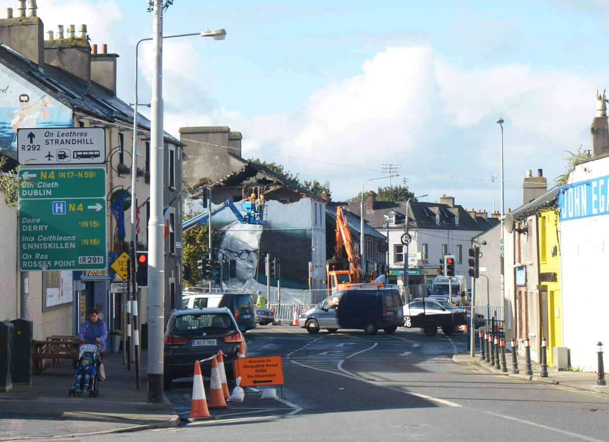 city streets in Sligo Ireland with houses, pedestrians and road. A crane is being used to demolish the top part of a house. On the side of that house, exterior, is a large mural that is a portrait of the poet William Butler Yeats (W.B. Yeats)