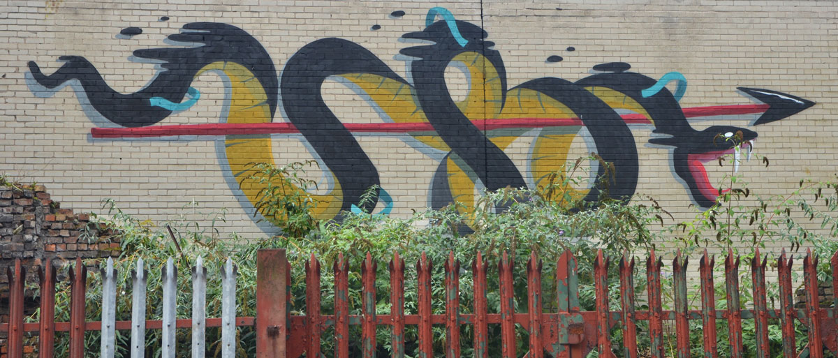 a rusty red fence and a green hedge is in front of a mural of a large black and yellow snake wrapped loosely around a long red arrow. 