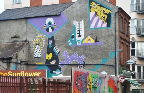 a collection of street art pieces on the side of a greybuilding, painted in purple, yellow and turquoise mostly. Abstract shapes