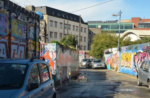 construction hoardings covered with street art and graffiti with cars parked beside them. 