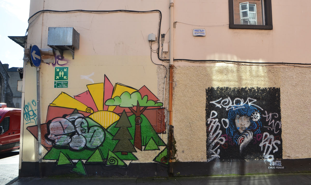 two street art paintings on a yellowish stucco wall, side of a building. One is a stylized landscape with trees, mountains and sun that has a large tag on top of it. The other is a person with long blue hair, hand by chin, head and shoulders only, by Evolve Urban Art
