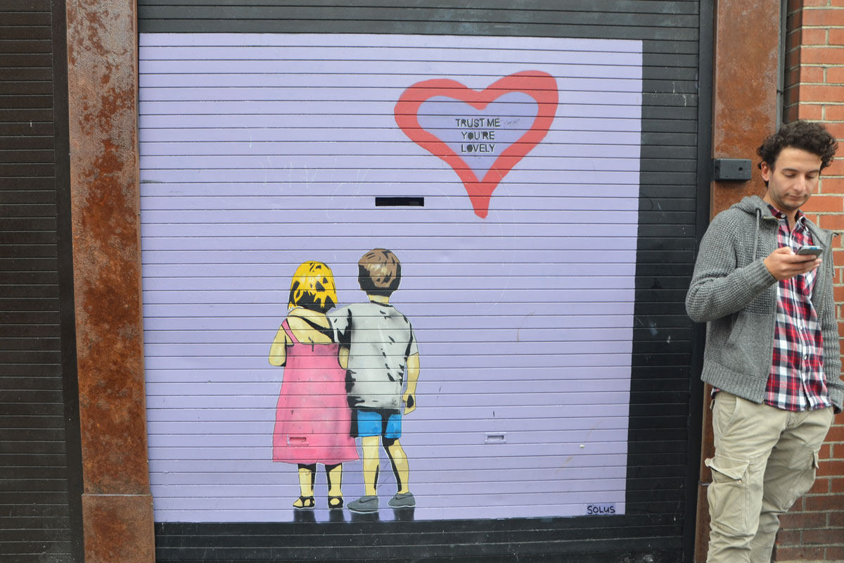 a young man on his phone is standing in front of a street art painting, lavender colour background with a boy and girl withtheir back to the viewer. THe boy has his arm around the shoulder of the girl. There is a heart in the upper right corner, outlined in red and containing the words Trust me you're lovely.