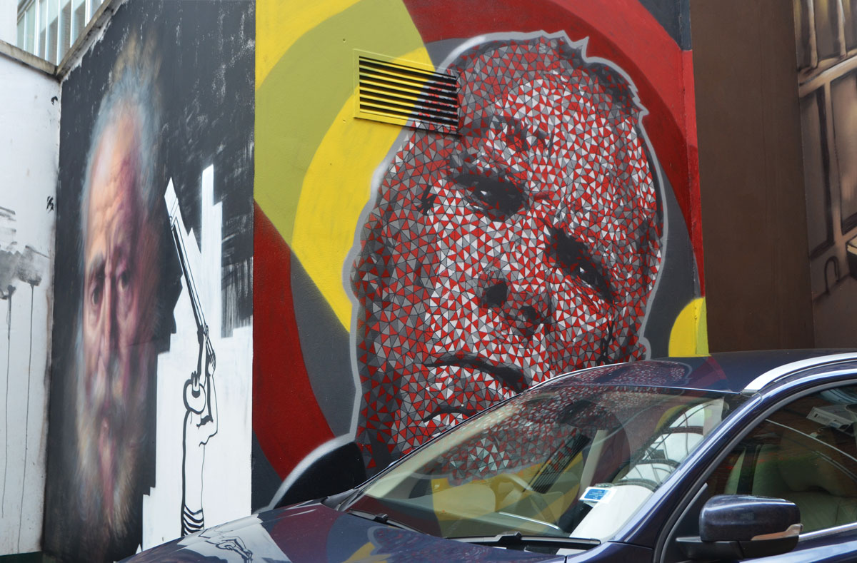two murals on two sides of a corner. One is a realistic painting of an old man and the other is also a man's head but it is done in red, white and black shapes. 