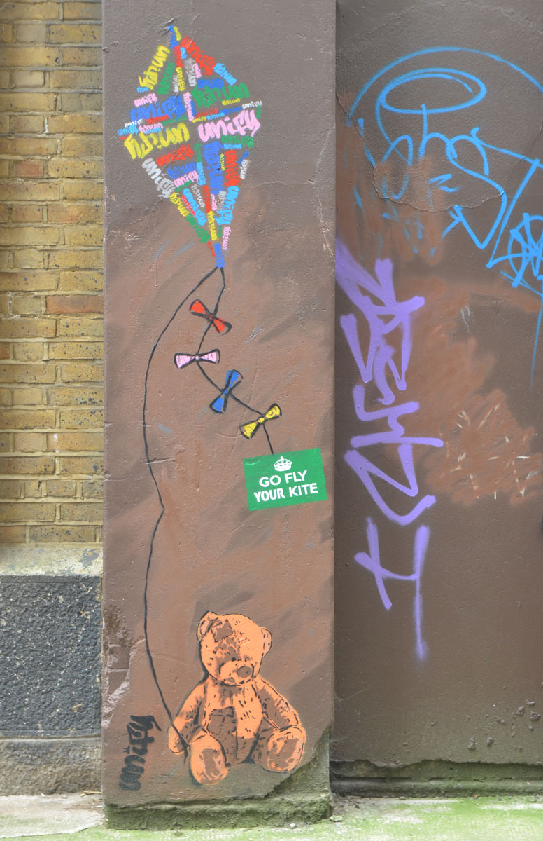 a teddy bear paste up or stencil sits on the sidewalk. he is holding a kite string in his paw and the kite is above him on the wall. A green sticker says go fly your kite. The signature says unify. the word unify is written many times in the kite in different colours to make a design on the kite. 