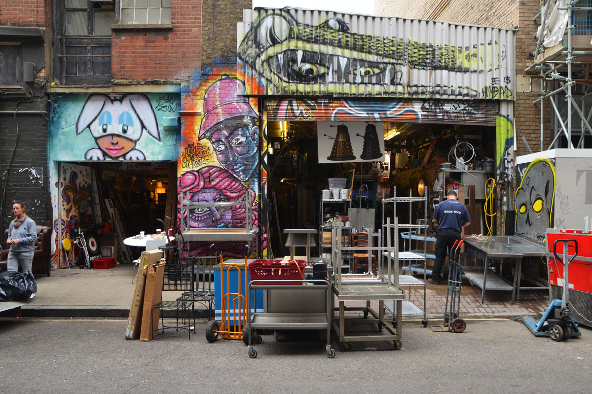 four or five pieces of street art beside and above a store front where there is a lot of activity going, moving items, delivering items. 