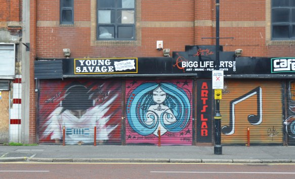 storefronts on a streeet in Belfast, brick building, but metal awnings/screens covering the shops because they are closed. Young Savage vintage clothing and Arts Lab, both have street art paintings on the screens. One is a young woman with long blue hair that forms a circle around her. 