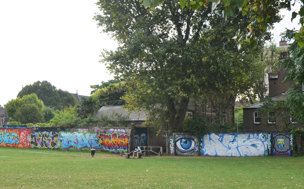 looking across a park, grass in the foreground. A man is sitting on a bench in front of fence covered with graffiti and street art, including a painting of a big blue eye 