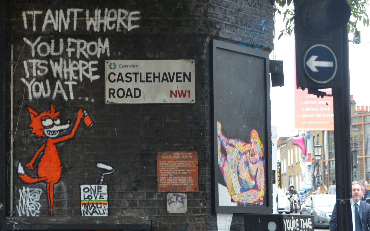 LOndon street sign, Castlehaven Road, Camden, NW1, with an orange cat street art piece beside it. The cat is spraying white words on the black wall, It ain't where you're from but where you are. 