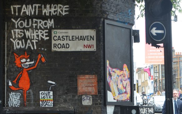 LOndon street sign, Castlehaven Road, Camden, NW1, with an orange cat street art piece beside it. The cat is spraying white words on the black wall, It ain't where you're from but where you are. 