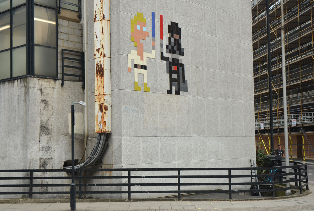 two large invader street artist pieces make of square tiles, a Luke Skywalker in white with a blue light saber and Darth Vader in black with his red light saber, on a grey concrete wall 