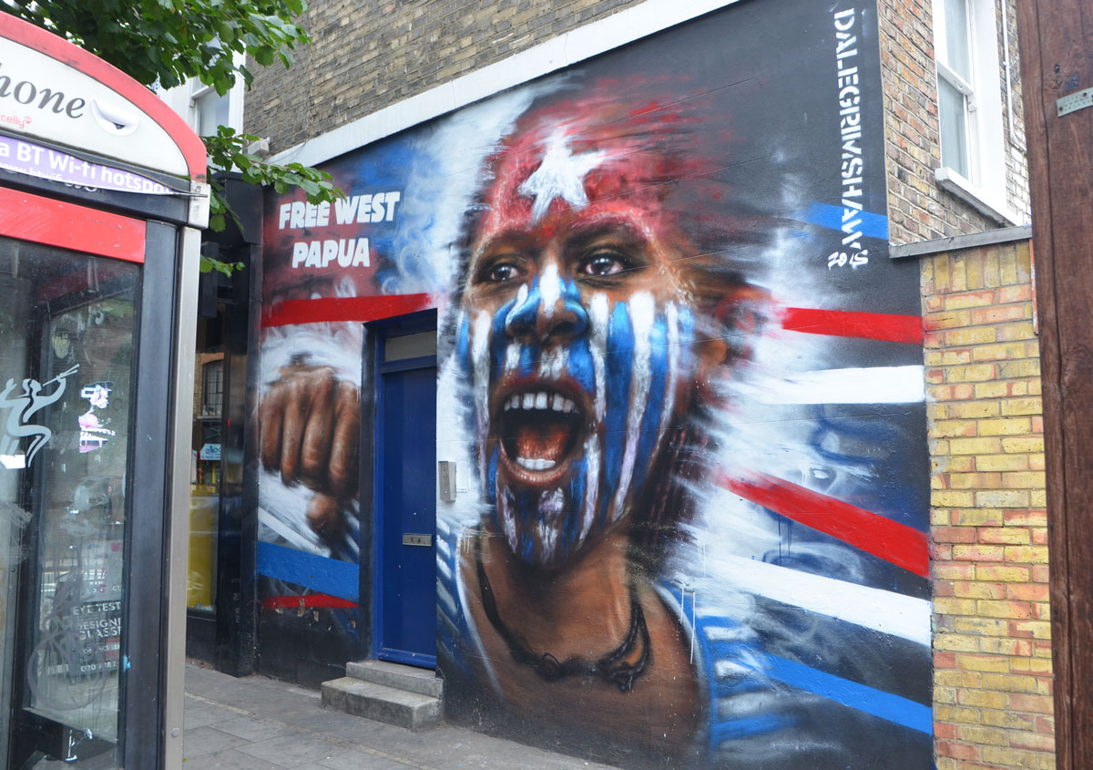 free west papua mural of a young aboriginal boy with a white star on red forehead and rest of face in blue and white stripes, by Dale Grimshaw, about 9 or 10 feet high, blue door in the wall in the mural