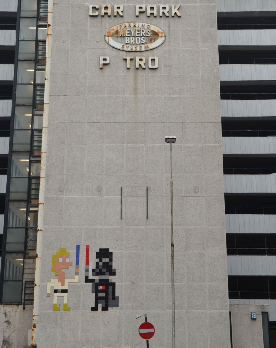 two large invader street artist pieces make of square tiles, a Luke Skywalker in white with a blue light saber and Darth Vader in black with his red light saber, on a grey concrete wall of the Meyers Parking building