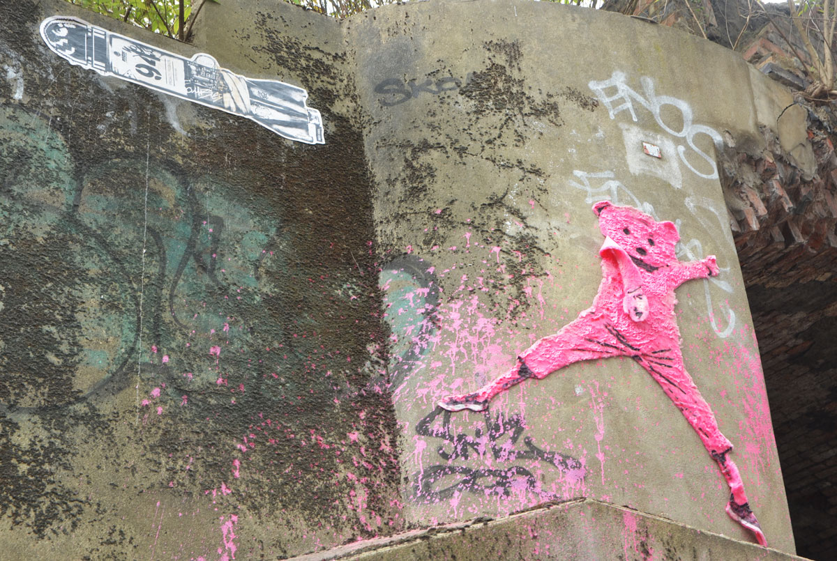 street art and graffiti in Shoreditch England, on Braithwaite Street, a pink teddy bear with terry cloth body high on a wall, beside a black and white pasteup of a bomb 
