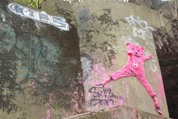 street art and graffiti in Shoreditch England, on Braithwaite Street, a pink teddy bear with terry cloth body high on a wall, beside a black and white pasteup of a bomb 