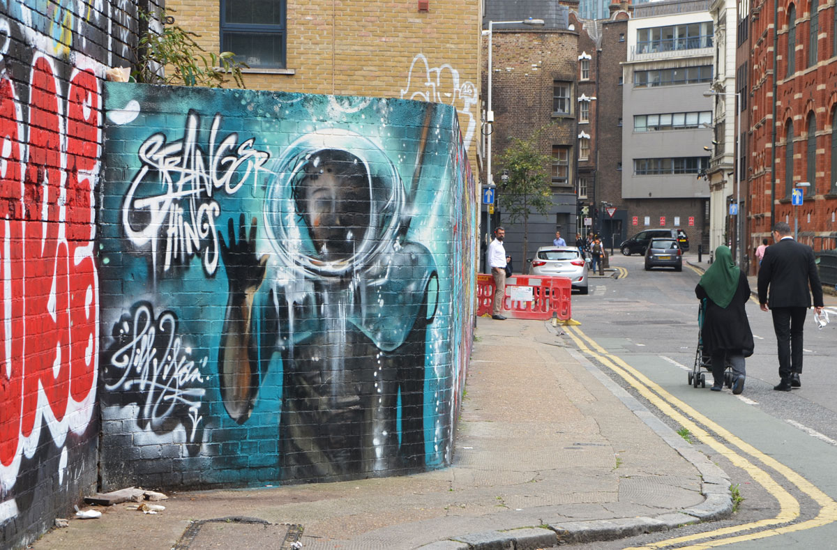 street art and graffiti in Shoreditch England, on Braithwaite Street, mural entitled strange things, man in old fashioned scuba mask waving, in turquoise background. 