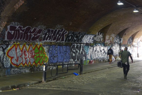 street art and graffiti in Shoreditch England, on Braithwaite Street, on the curved walls of an underpass, darkish, with a couple of people walking under the bridge