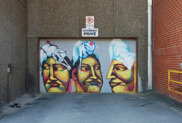 On a garage door, three heads, each has a cat on top. On of the cats has a mouse pulling a small red cart. 