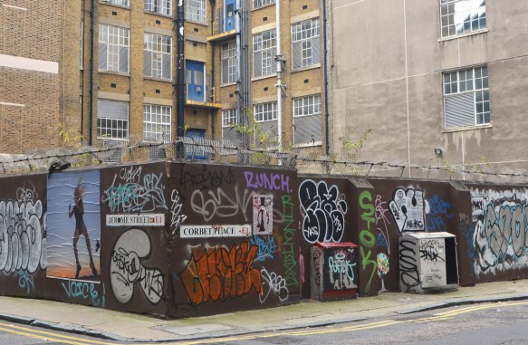 a wall covered with tags and other graffiti, stencils, paint, paste ups, surrounding a yard by an apartment complex, brick building in the background 