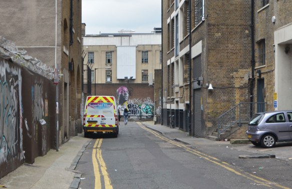 a truck painted in yellow and red stripes is parked on the side of a narrow street, brick three storey buildings on either side, graffiti on the wall at the end of the lane 