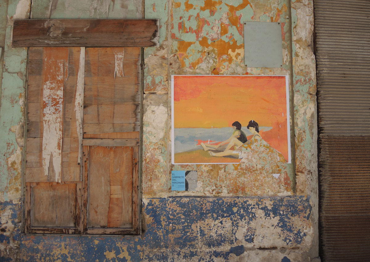 a rough wooden cover covers a window, a wall used to be orange and blue but most of the paint has peeled off. On the wall a picture has been mounted. It is a beach scene, two people sitting on an orange beach with orange sky and blue water. 