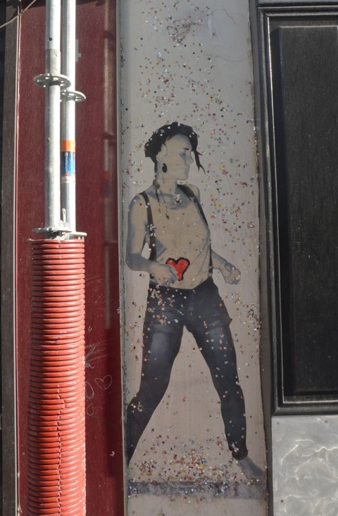 large size paper paste up of a young woman, standing, with a lot of small glittery pieces of paper scattered around her - wearing blue jeans and a white t-shirt with a red heart on it 