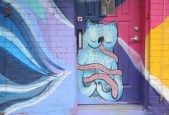 street art, graffiti, animals painted on a wall - blue cat with a very long pink tongue that encircles its body a couple of times, sitting in a doorway