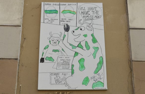 a poster showing a cow in a fast food restaurant ordering a human meat burger. Another cow is behind the cash register. Pictures of items on the menu are above the cashier. Done in cartoon style 