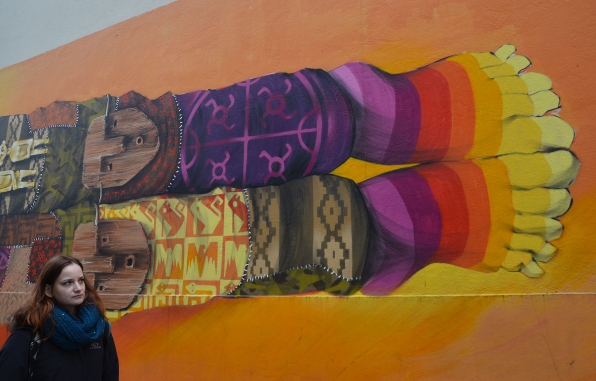 the end of a hotizontal mural of man bedecked in Inca symbols and colours, wood masks on his knees, patterned knit socks with symbols, not matching, rainbow coloured socks with toes. 
