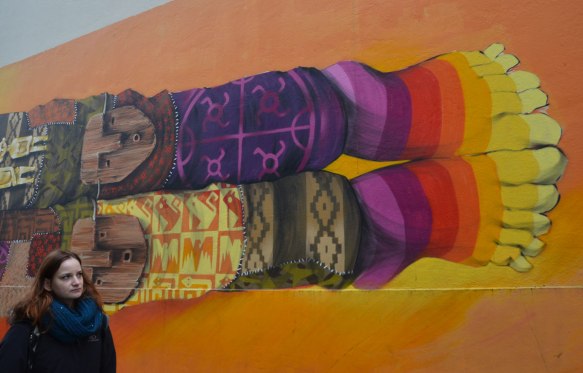 the end of a hotizontal mural of man bedecked in Inca symbols and colours, wood masks on his knees, patterned knit socks with symbols, not matching, rainbow coloured socks with toes. 