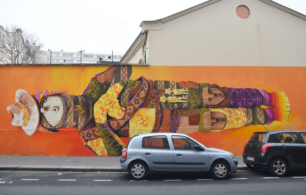 man in a long horizontal mural, cloth puppet like face, dressed in a patchwork of colours and patterns and Inca and South American symbols, two gold good luck charms attached to his belt. 