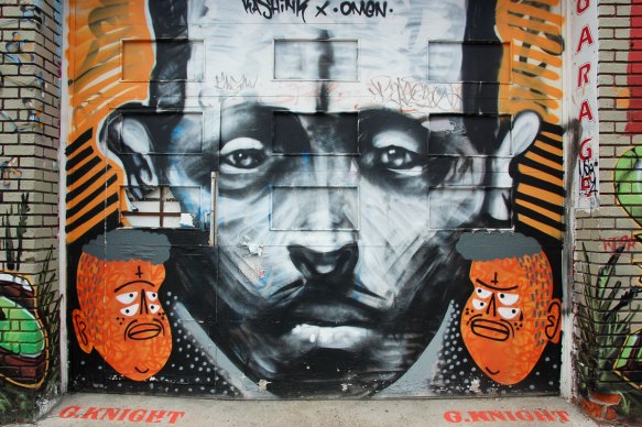 large black and white man's face painted by omen with a small orange face to the bottom left and bottom right - orange faces painted by kashink