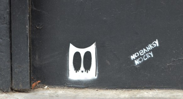 on a black wall, two small stencils, one is a white ghost head with big black oval eyes and the other is the words: no banksy no cry