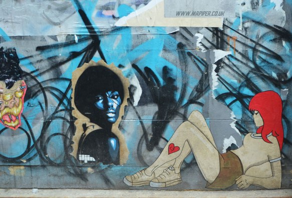 3 paste ups of women on a wall. One is a woman's head and shoulders in blue with short black hair by iljin . One is a multicoloured face. One is a young woman sitting on the ground with her knees up. She has red hair and a heart on her leg, by c3b