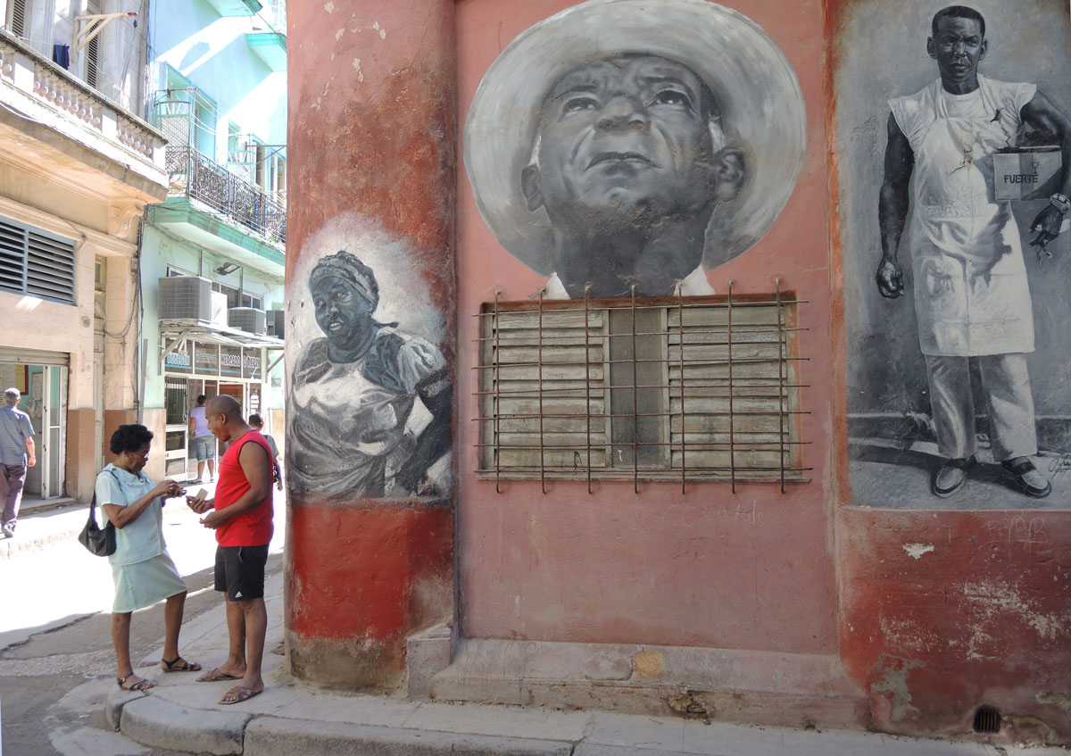 a man and a woman talking on a street corner. Beside them is a wall with three black and white murals painted on it. A middle ages woman with a head band and hands on her hips, an older man's head in a white hat and looking upward and last, a man in a white uniform holding a box under his arm. 