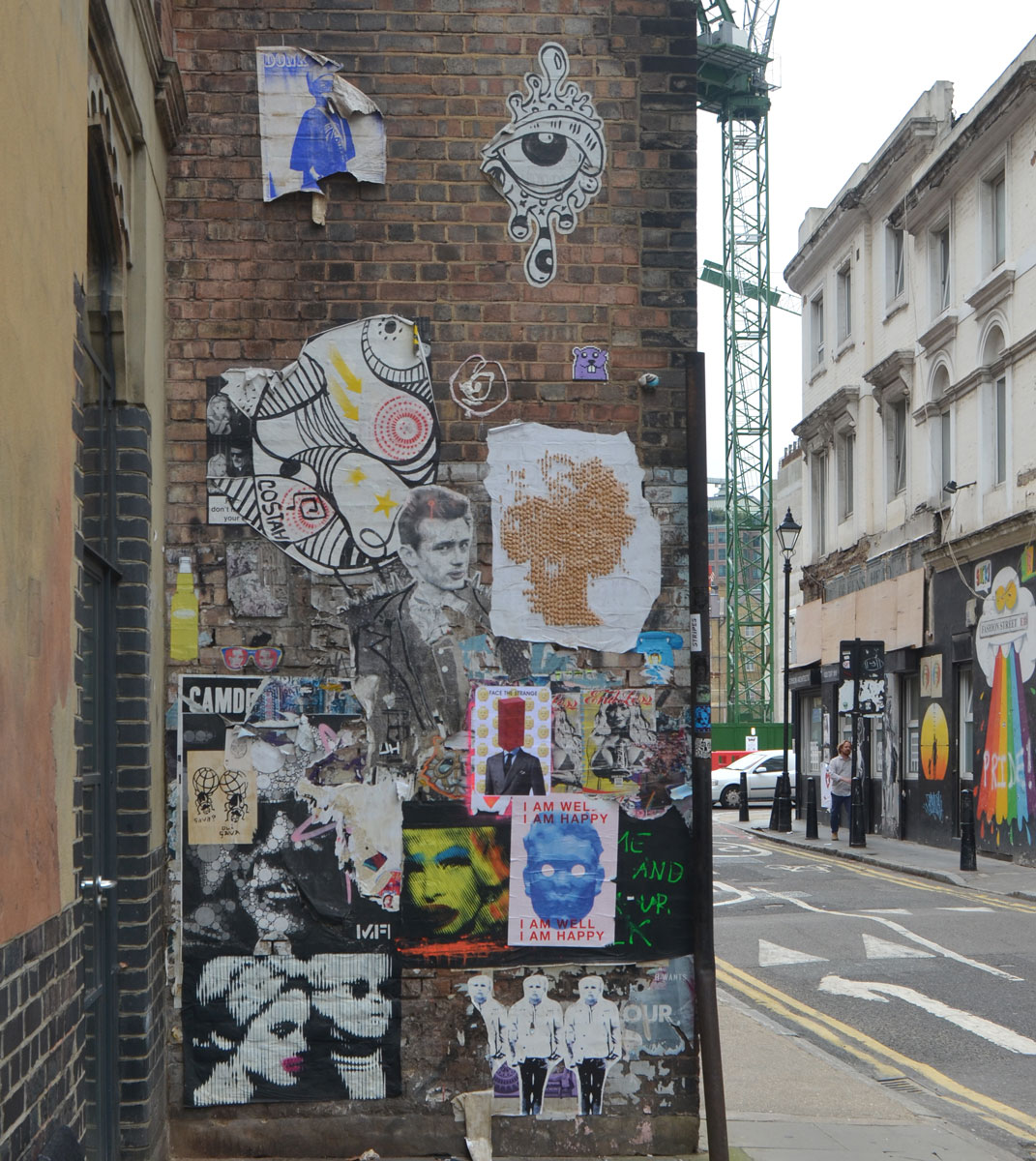 a brick wall on Fashion Street in Shoreditch that is covered with a lot of paste up pieces of graffiti and street art 