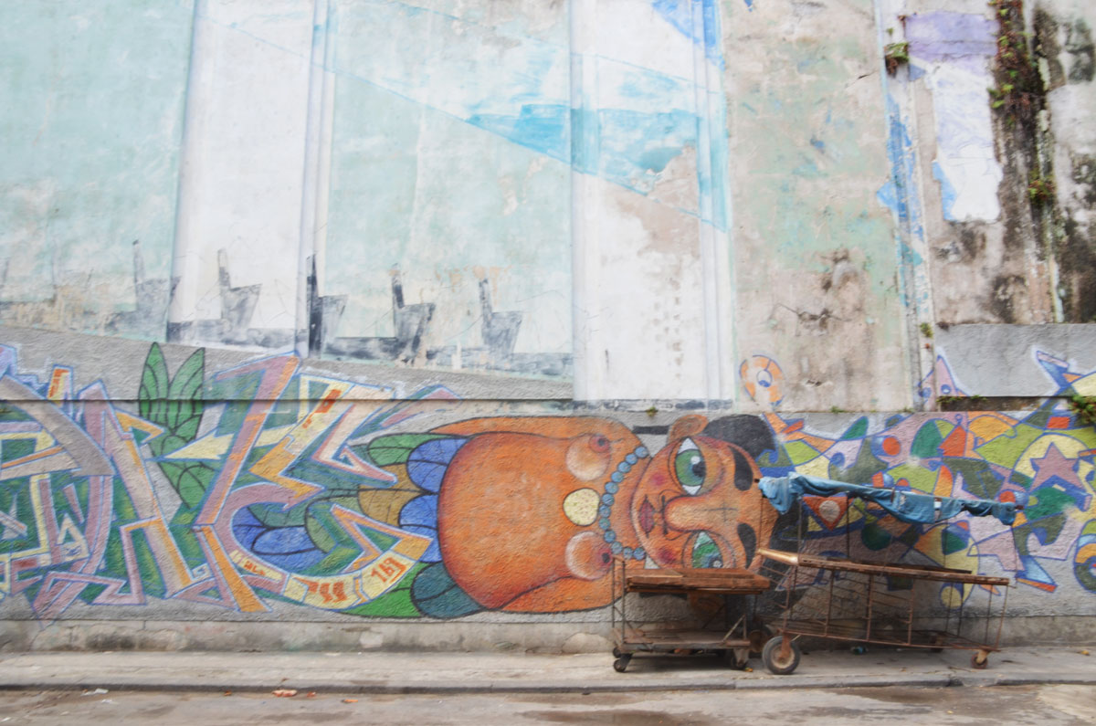 a wooden cart in poor shape is parked next to a mural of a woman lying on her side. It looks like she is lying on the sidewalk. She is topless. 