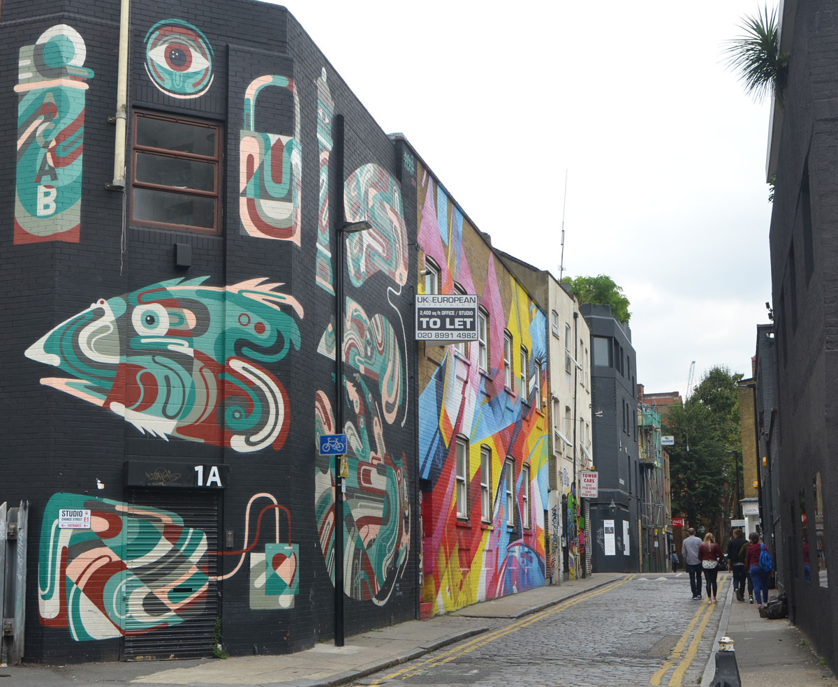 looking down Chance Street in Shoreditch, a few people walking on the narrow street, the buildings on the left are covered with murals. The first is aboriginal symbols in white, green and brown on a black background. THe second is in bright shades of blue, red and yellow. 
