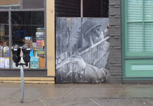 black and white photo of a man playing the flute, pasted onto a woodedn fence beside a sidewalk, next to a book store, a parking meter nearby, on a street in Buffalo 