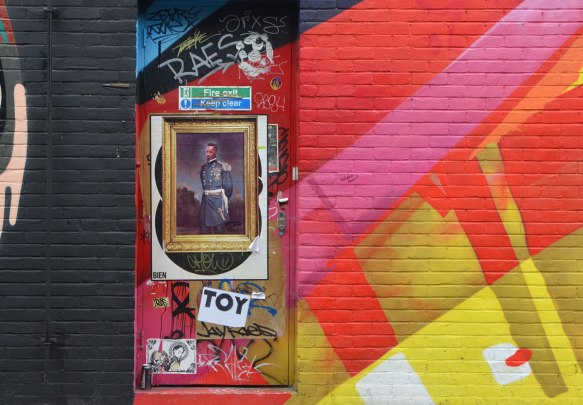 a door with pasteups on it, between a black wall and a wall that is painted in red, pink and yellow. One of the paste ups is of a black man in a military uniform in a gilded frame. The word rai is written on top of it. 