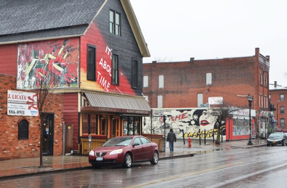 a brick two storey store with a mural on the upper part in red, yellow and black, with the words It's about time. The picture in the mural is three red fists. One is holding a yellow paint roller and one is holding a yellow spray paint can. Another mural is in the background, a car parked on the wet street, a pedestrian on the sidewalk 