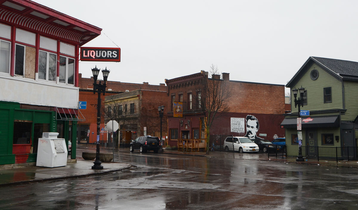 intersection in Buffalo with old two storey square front storefronts. wet road, rainy day, liquor store on the corner that is now empty but the sign only half works - only half the letters in the word liquor are lit. 