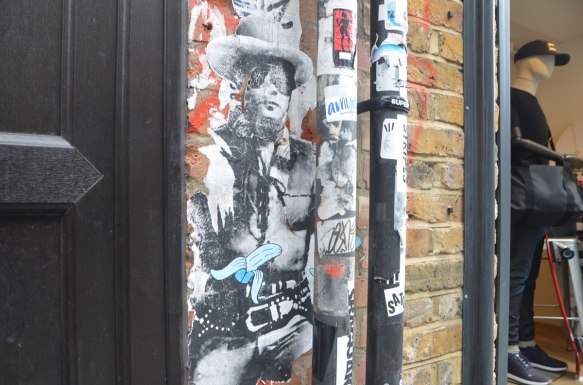 a pasteup of a black and white print of a cowboy, not quite life sized, with cowboy hat and vest. The paper is starting to peel away at the edges. A sticker of a blue and white partially peeled banana has been stuck on him. Beside him is a male mannequin in a store window. 