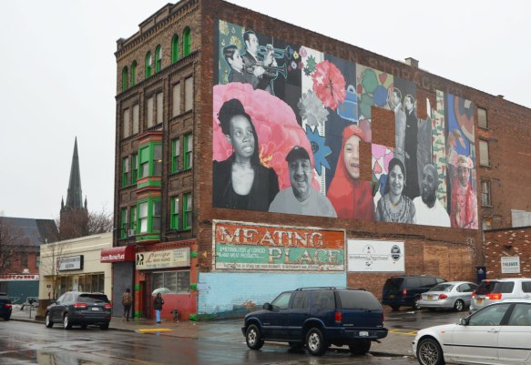 5 storey brick building on Grant St in Buffalo, with green trim around the windows and a small parking lot beside, sign on the side of the building says Meating Place. There is a large mural on the side of the building featuring realistic portraits of many people, including two trumpet players, a girl in a red head scarf, and other men and women, 